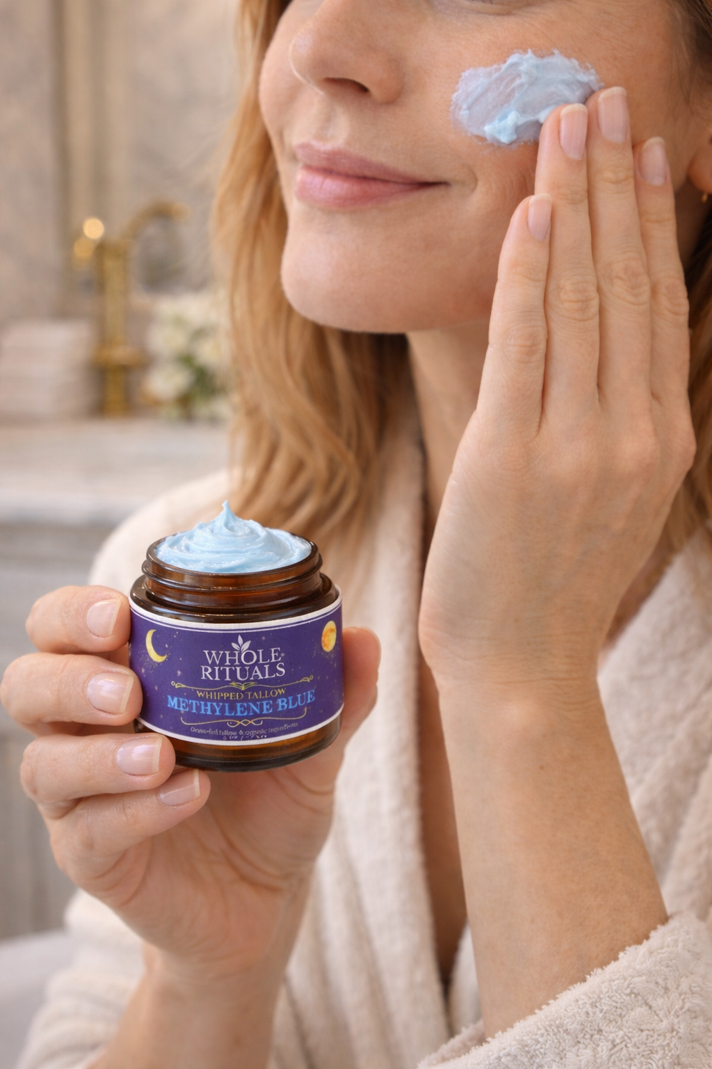 Woman applying a blue cream to her face with a jar of the same cream in her hand.