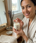 Woman in a bathrobe holding a jar of body scrub in a home setting.